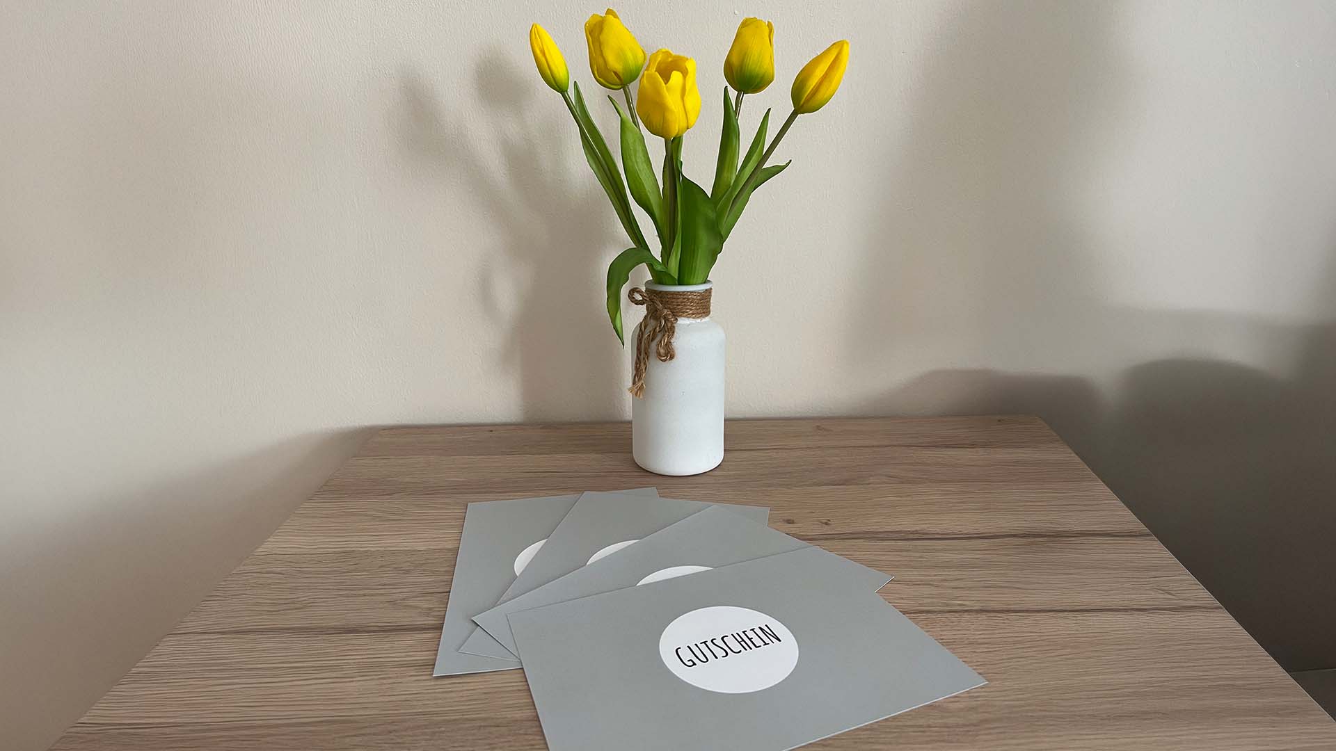 Auf einem kleinen Holztisch liegen die Gutscheine für Sitzungen bei Manuela. Im Hintergrund ist ein Blumenstrauß zu sehen mit gelben Tulpen.