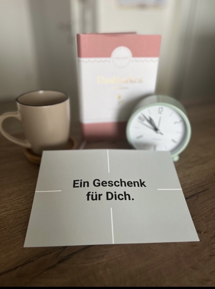 Auf einem Tisch stehen: - ein Geschenkbuch - ein grüner Wecker - eine beige Tasse - ein Gutschein, der mittig im Bild ist