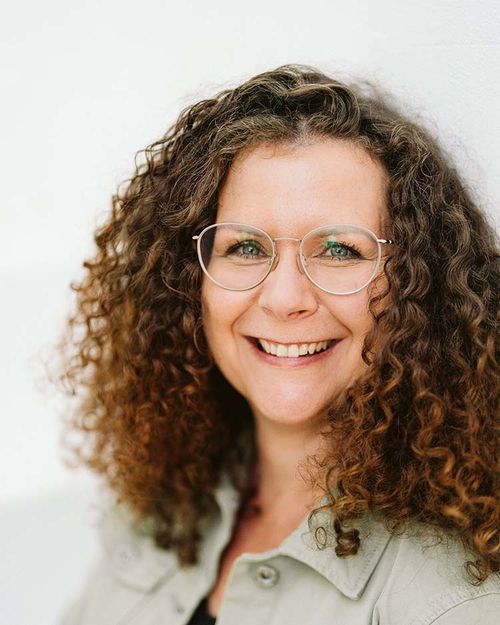 Portrait von Manuela Herold. Sie trägt eine leicht grüne Jeansjacke, eine Brille und hat lockige Haare.