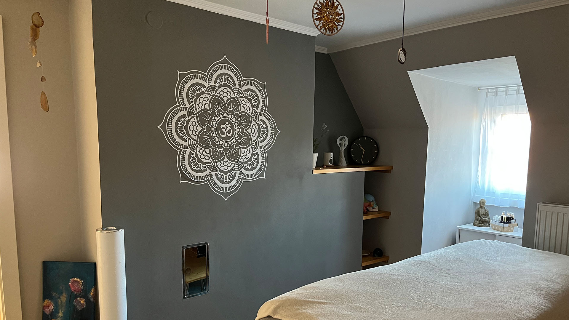 Bild der Praxis: Rechts unten steht eine Liege, links ist eine dunkellackierte Wand mit einem Wandtattoo, welches an den Hinduismus erinnert.